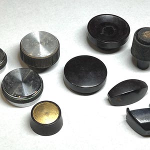 May include: A collection of vintage knobs in various shapes and sizes. The knobs are made of metal and black plastic, with some featuring a textured or ridged design. The assortment includes round, oval, and hexagonal shapes, suitable for electronics or vintage equipment.