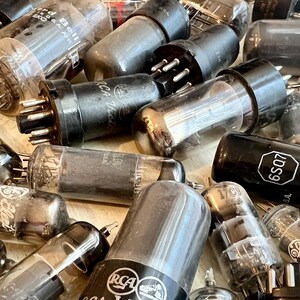 May include: A collection of vintage vacuum tubes, featuring glass and black cylindrical components with metal pins. Some tubes bear the RCA logo and model numbers like 6SQ7. The tubes are arranged in a close-up, slightly angled view.