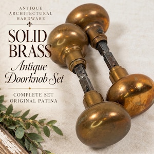 May include: A set of two round solid brass doorknobs with a dark metallic patina, connected by a square metal spindle. The text on the image reads: ANTIQUE ARCHITECTURAL HARDWARE, SOLID BRASS, Antique Doorknob Set, COMPLETE SET ORIGINAL PATINA.