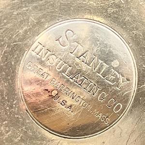 May include: Close-up of a silver-toned metal object with the embossed text "STANLEY INSULATING CO. GREAT BARRINGTON, MASS. U.S.A." The metal surface shows wear and scratches, with the text in a circular design.
