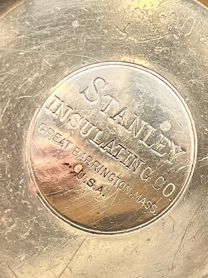 May include: Close-up of a silver-toned metal object with the embossed text "STANLEY INSULATING CO. GREAT BARRINGTON, MASS. U.S.A." The metal surface shows wear and scratches, with the text in a circular design.