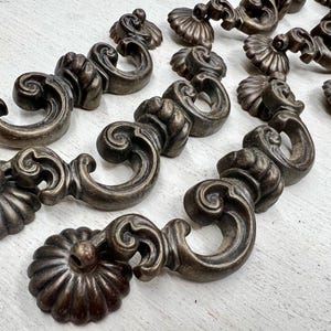 May include: A collection of antique bronze-toned drawer pulls with a decorative, ornate design. Each pull features a curved handle with a scroll design and a rosette-shaped base. The hardware is arranged on a light-colored surface.
