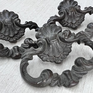May include: Close-up of four antique bronze drawer pulls. Each pull features a decorative floral design with a central square element and a curved handle. The pulls are arranged on a white surface.