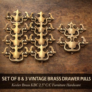 May include: A collection of vintage brass drawer pulls arranged on a wooden surface. The pulls have a decorative design with curved handles and ornate detailing. The text at the bottom reads "SET OF 8 & 3 VINTAGE BRASS DRAWER PULLS" and "Keeler Brass KBC 2.5" C/C Furniture Hardware."
