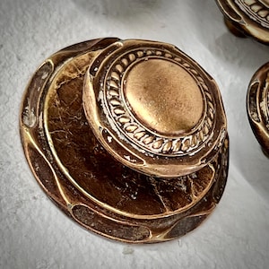 May include: Close-up of antique brass door knobs. The knobs have a circular design with a central, raised circle surrounded by a decorative border. The outer edge features an ornate, layered design with a distressed finish.