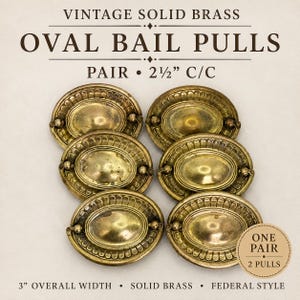 May include: A set of six vintage solid brass oval bail pulls with a 2.5-inch center-to-center measurement and a 3-inch overall width. The hardware features a decorative fluted border around the central oval plate and a hinged handle.