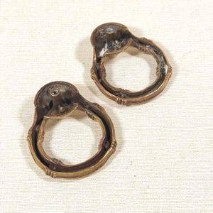 May include: Two circular brass drawer pulls with a weathered patina. Each pull features a hinged ring attached to a mounting plate with a central screw hole. The metal surfaces show signs of age and oxidation, with a textured finish.