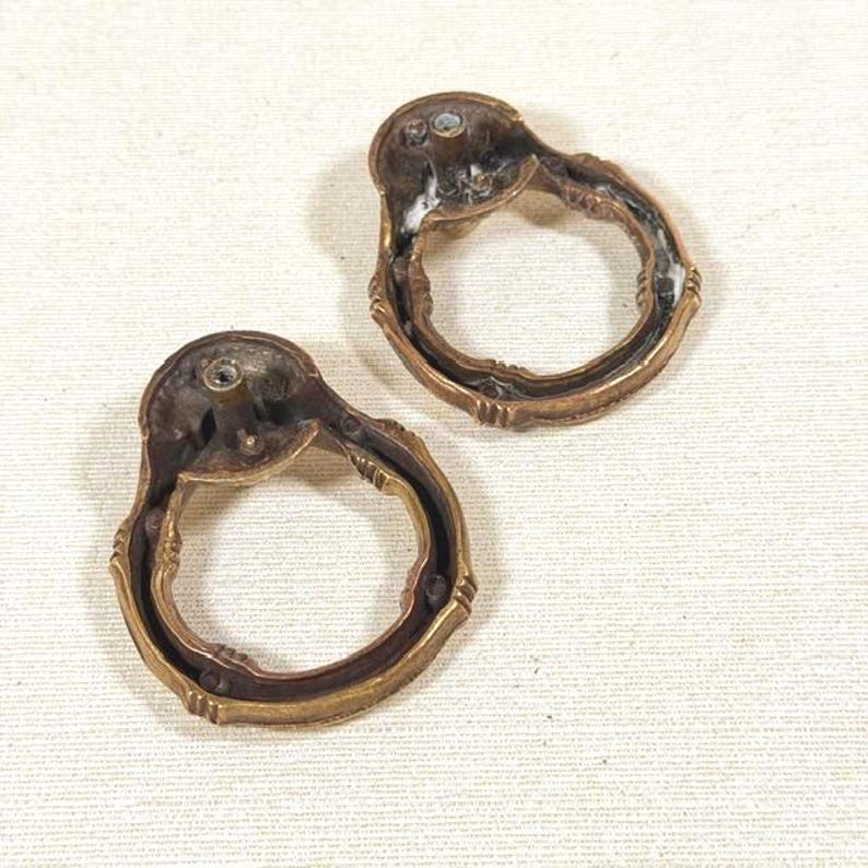 May include: Two circular brass drawer pulls with a weathered patina. Each pull features a hinged ring attached to a mounting plate with a central screw hole. The metal surfaces show signs of age and oxidation, with a textured finish.