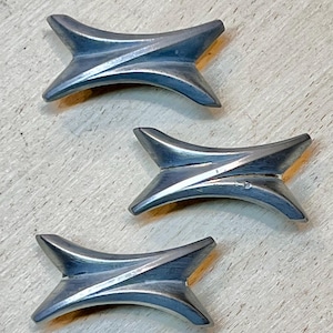 May include: Three silver-toned, star-shaped objects with curved edges are arranged on a light-colored surface. Each object has a central groove and pointed ends, suggesting a decorative or functional design.