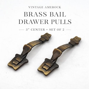 May include: Two vintage brass bail drawer pulls with a 3-inch center. The pulls have a curved handle and a rectangular base with decorative detailing. The text "VINTAGE AMEROCK" and "BRASS BAIL DRAWER PULLS" are visible.