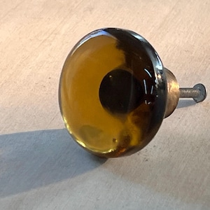 May include: A round cabinet knob with a clear amber glass dome and a dark brown center. The knob has a brass base and a silver screw for attachment. This decorative hardware is suitable for drawers and cabinets.