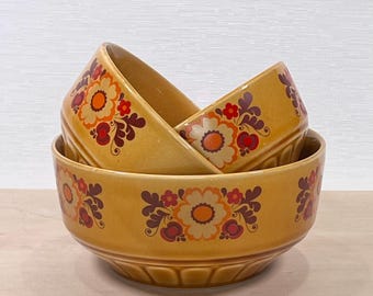 May include: Three stacked yellow ceramic bowls with a floral pattern in orange, red, and purple. The bowls have a scalloped edge and are nested inside each other. The floral design is repeated around the bowls.