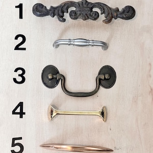 May include: Five different cabinet hardware pulls in various styles and finishes. The pulls include a dark, ornate cast iron pull, a brushed nickel pull, a bronze pull with a handle, a brass pull, and a copper pull.