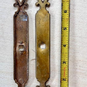 May include: Two antique brass door pulls with ornate, symmetrical designs. One is tarnished, the other is polished. A yellow measuring tape shows the pulls are approximately 7 inches long.