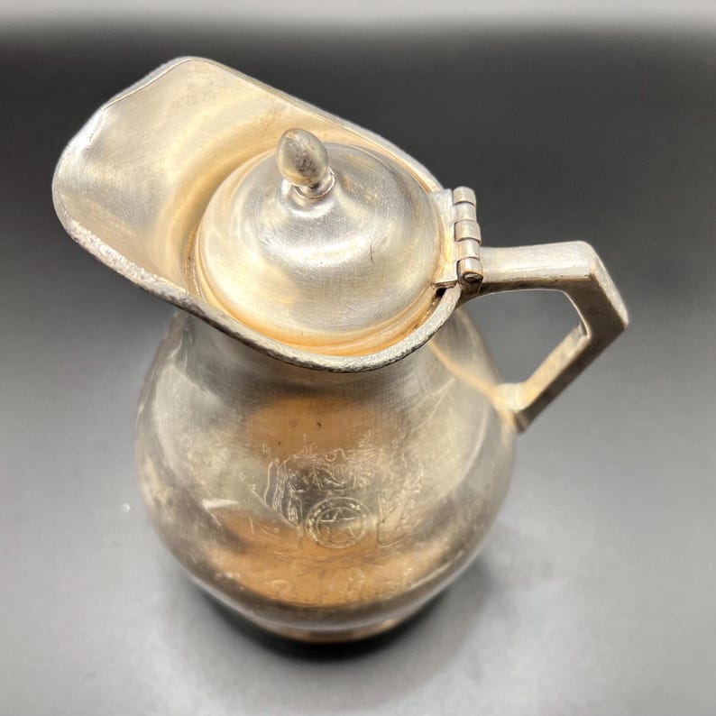 May include: A small, silver-colored pitcher with a hinged lid and a handle. The pitcher has a rounded body and a spout. The lid has a small knob on top. The pitcher appears to be made of metal and has a vintage look.