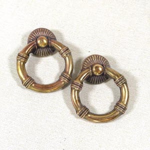 May include: Two circular brass drawer pulls featuring a hinged ring design. Each pull has a decorative fan-shaped backplate with a central knob and a textured ring with three distinct ribbed sections along the circumference.