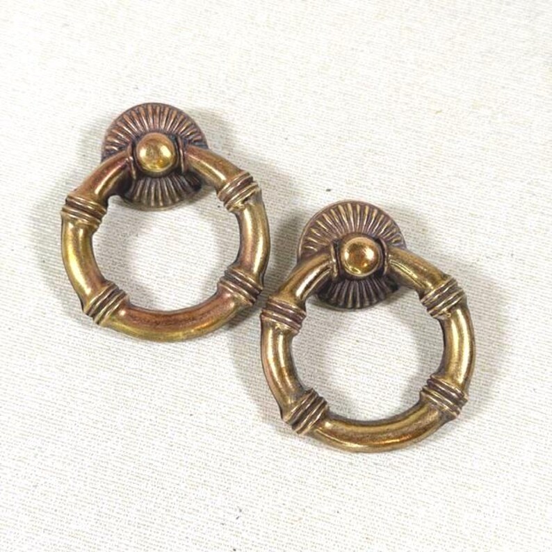 May include: Two circular brass drawer pulls featuring a hinged ring design. Each pull has a decorative fan-shaped backplate with a central knob and a textured ring with three distinct ribbed sections along the circumference.