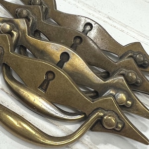 May include: A collection of antique brass drawer pulls. Each pull features a keyhole and a decorative design with pointed ends and a rounded handle. The pulls have a weathered, aged appearance.