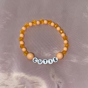 Op de afbeelding: Een armband met perzik- en oranje kralen. Het woord "CUTIE" is gespeld met witte letterkralen. De armband ligt op een roze ondergrond.