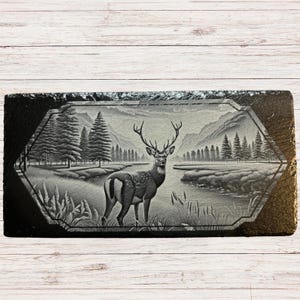 May include: A black and white engraved slate plaque featuring a deer standing in a forest scene. The deer has antlers and is looking to the right. The background shows trees, mountains, and a river.