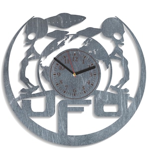 Puede incluir: Un reloj de pared redondo de color plateado con textura de madera. La esfera del reloj tiene una aguja de hora negra y una aguja de segundos roja. La esfera del reloj está rodeada por un diseño de dos extraterrestres con un OVNI sobre ellos. La esfera del reloj también tiene la palabra "UFO" impresa debajo.