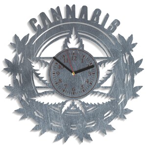 Op de afbeelding: Een zilveren metalen wandklok met een cannabisbladontwerp en het woord "Cannabis" geschreven rond de buitenrand. De wijzerplaat van de klok heeft zwarte wijzers en bruine cijfers.