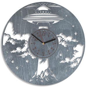 May include: A gray wooden clock with a cut-out design of a UFO flying over clouds and mountains. The clock face has black hands and a red second hand.