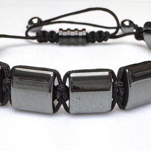 May include: A dark gray hematite bracelet with rectangular beads strung on a black cord. The bracelet has a sliding knot closure for adjustable sizing. The beads are polished and reflect light.