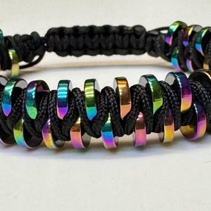 May include: A braided bracelet featuring iridescent metal rings threaded onto black cord. The rings display a spectrum of colors, including purple, green, and gold. The bracelet is designed with a macrame style and is suitable for everyday wear.