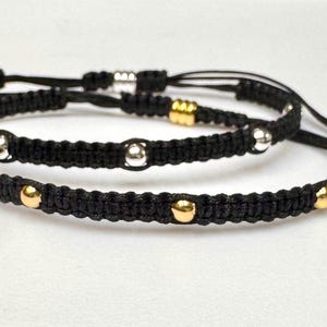 May include: Three black macrame bracelets with adjustable sliding knots. Each bracelet features metallic silver and gold-colored beads. The bracelets are displayed on a white surface.