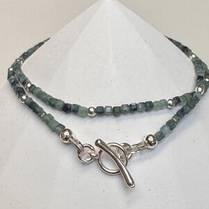 May include: A silver chain necklace with a toggle clasp. The chain is made of small, square-shaped green gemstones.