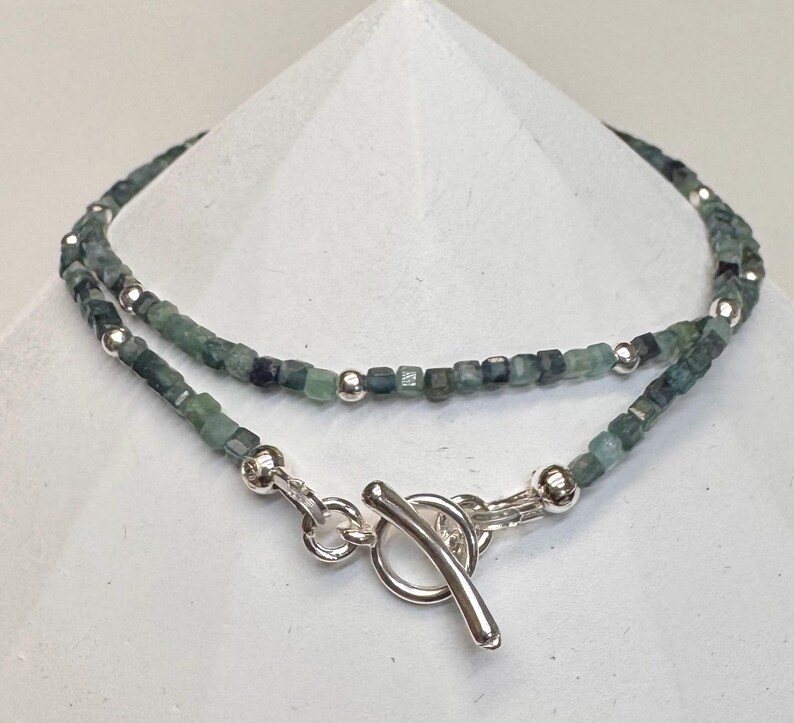 May include: A silver chain necklace with a toggle clasp. The chain is made of small, square-shaped green gemstones.