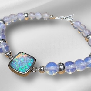 May include: A bracelet featuring iridescent opal and pale blue beads. The square opal is set in a gold-colored bezel. The bracelet has silver-colored accents and a toggle clasp.
