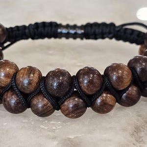May include: A double-strand bracelet featuring dark brown wooden beads. The beads are strung on black cord, with adjustable black end beads. The bracelet has a macrame style design and a natural, earthy aesthetic.