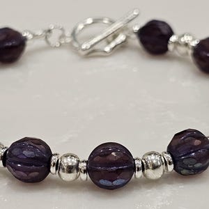May include: A beaded bracelet featuring dark purple, faceted beads and silver-colored accent beads. The bracelet has a toggle clasp closure. The beads are strung on a flexible cord.