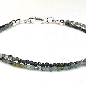 May include: A delicate bracelet featuring two strands of small, faceted beads in shades of green, blue, and gray. The bracelet is secured with a silver-colored clasp. The jewelry is displayed on a white background.