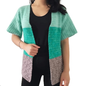 Peut inclure: Cardigan en crochet à manches courtes et ouvert sur le devant, dans des tons vert menthe, turquoise et gris. Le design en blocs de couleurs présente un haut vert menthe, une section centrale turquoise et un bas gris. Fait main, coupe décontractée.