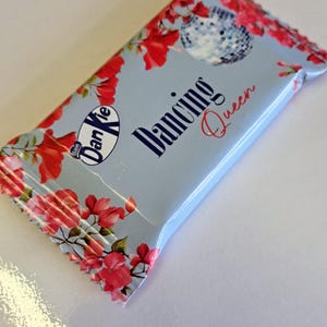May include: A blue and white candy wrapper with a pink floral design. The wrapper has a disco ball and the text "Dancing Queen" on it. The wrapper is for a candy bar made by "Babo Dan Kie".