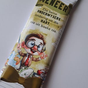 Op de afbeelding: Een bruine en witte chocolade reep met een cartoon illustratie van een leraar met een strikje en bril. De tekst op de reep luidt "Meneer Die beste ONDERWYSERS onderrig uit die HART, nie uit boeke nie."