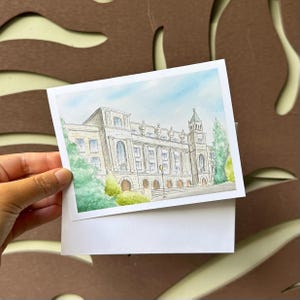 May include: Watercolor painting of a stately building with a light blue sky background. The building is white with arched windows, a tower, and green trees. The painting is on a white card.