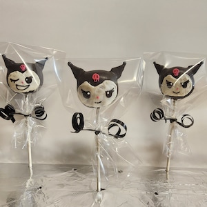 May include: Three Kuromi-themed cake pops. Each cake pop features a black and white face with a pink skull and black ears. They are individually wrapped in clear cellophane and tied with black ribbon on a stick.