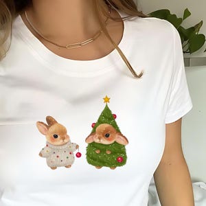 May include: White t-shirt featuring two adorable bunny illustrations. One bunny wears a sweater and holds a red ornament. The other bunny is inside a Christmas tree. A gold necklace is visible.