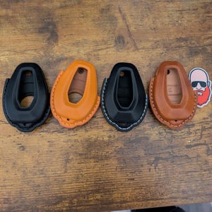 May include: Four key fob covers in different colors and materials. Two are black leather with white stitching, one is orange leather with orange stitching, and one is brown leather with brown stitching.