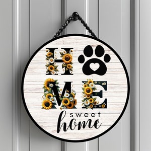 May include: Round wooden sign with the word "HOME" spelled out in black letters with sunflowers and white daisies around each letter. A black paw print with a heart in the center is between the "H" and the "O". The words "sweet home" are written in black cursive below the letters.