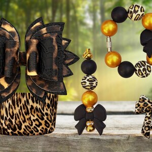 May include: A set of accessories featuring a large leopard print bow, a leopard print cuff, a beaded bracelet with gold, black, and leopard print beads and a black bow, a beaded keychain with a black bow, and a small leopard print bow hair clip.