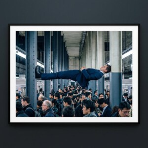 May include: A framed print depicts a man in a blue suit seemingly levitating in a crowded subway station. He is leaning against a metal support beam, with a sea of people below. The image has a surreal, dreamlike quality.