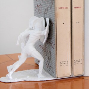 May include: White decorative bookend featuring a classical warrior figure with a shield and helmet. The bookend is positioned next to two vintage books with the title "Germinal" and the publisher "Andre Sauret Editeur".