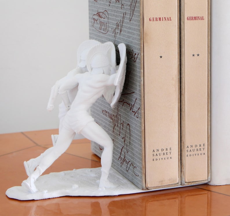 May include: White decorative bookend featuring a classical warrior figure with a shield and helmet. The bookend is positioned next to two vintage books with the title "Germinal" and the publisher "Andre Sauret Editeur".