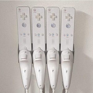 Può includere: Un supporto da parete bianco con quattro controller per videogiochi bianchi e quattro telecomandi bianchi attaccati ad esso.