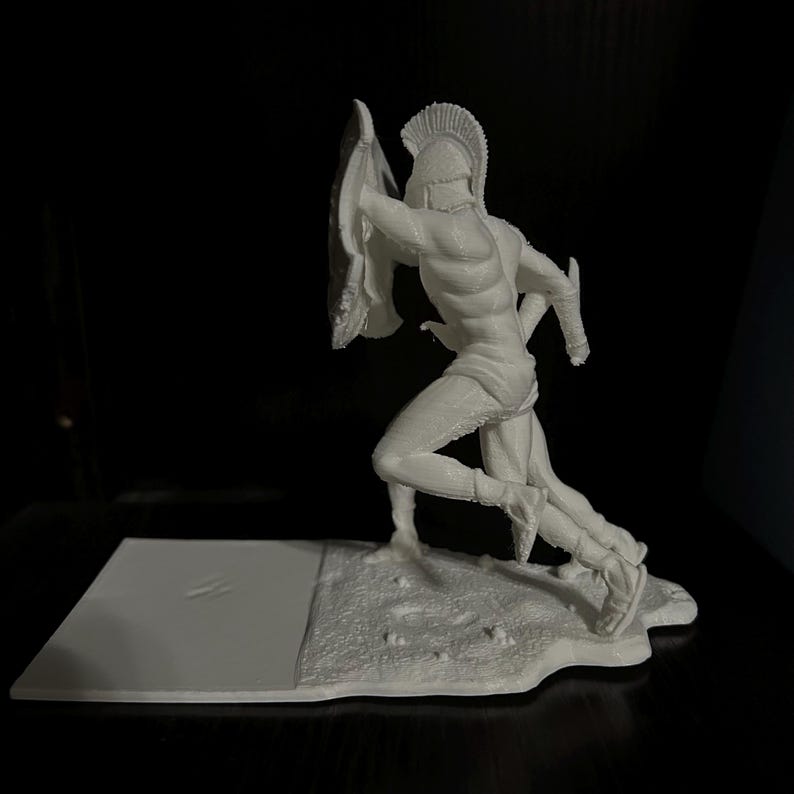 May include: A white figurine of a running Spartan warrior, complete with a helmet, shield, and sword. The figure is posed mid-stride, with detailed musculature and armor. The base is a textured, irregular shape.